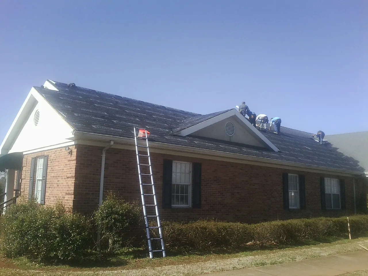 Crew tearing shingles off a brick ranch roof with a ladder set — Metal Roof Repair work by our crew in Wesley