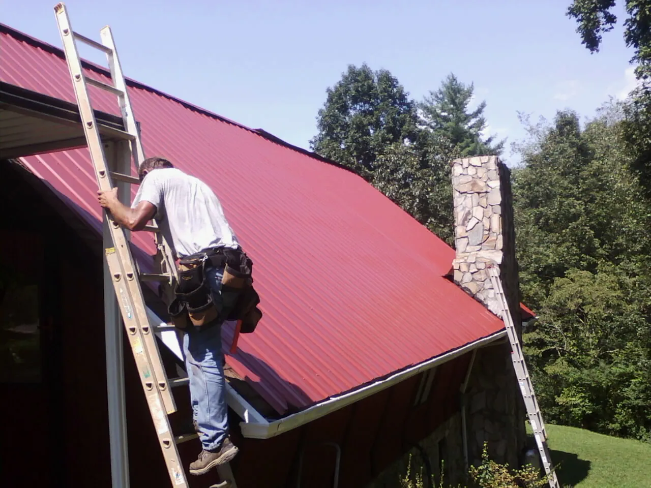 Roofer on a ladder beside a red metal roof and stone chimney — Soffit & Fascia Repair by Roofing Brothers in Wesley
